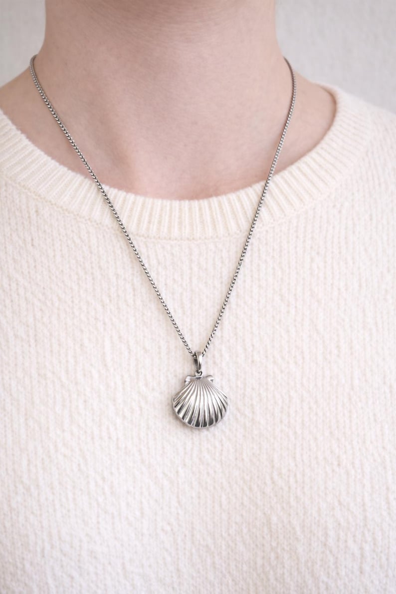 May include: A silver seashell pendant necklace. The pendant is a detailed, three-dimensional scallop shell design. The necklace chain is a delicate, linked style. The person is wearing a cream-colored sweater.