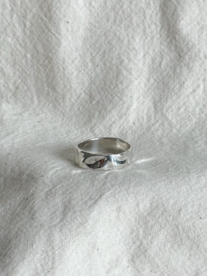 May include: A silver ring with a textured, uneven surface. The ring is wide and has a smooth, polished finish. The ring is set against a white, textured background.