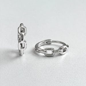 Pode incluir: Dois brincos de argola prateados com elos de corrente. Um brinco está em pé, o outro está plano. Os brincos são feitos de metal brilhante e estão sobre um fundo branco. O design é simples e moderno.