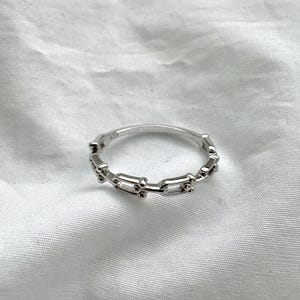 May include: A silver ring with a chain link design. The ring is made of a shiny, reflective metal and features a series of rectangular and circular links. The ring is set against a white background.