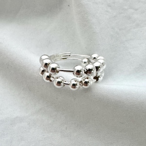 May include: A pair of silver hoop earrings. Each earring features a series of small, shiny, silver spheres strung along the hoop. The earrings are displayed on a white, textured surface, creating a simple, elegant presentation. The design is modern and minimalist.