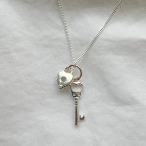 May include: A silver necklace featuring a heart-shaped locket with a keyhole and a small key charm. The necklace hangs on a delicate chain against a white background. The key charm has a detailed design.