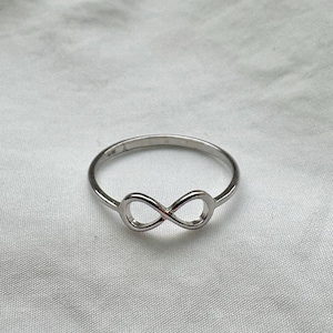 May include: A silver ring with an infinity symbol design. The ring is set against a white, textured backdrop. The infinity symbol is the focal point, with a smooth, polished finish.