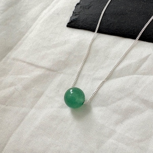 May include: A delicate silver necklace featuring a vibrant green, spherical gemstone pendant. The gemstone is a smooth, polished sphere, suspended from a fine silver chain. The necklace rests on a white fabric surface, with a dark, textured background.