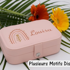 Peut inclure: Une boîte à bijoux rose avec le nom "Louisa" et un motif arc-en-ciel. La boîte est rectangulaire avec une fermeture à boucle. Le fond comprend des fleurs orange et jaunes et des feuilles vertes. Le texte "Plusieurs Motifs Disponibles" est en bas.
