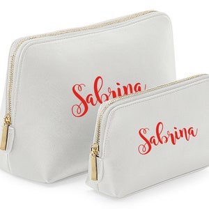 May include: Two white zippered cosmetic bags with gold zippers. The larger bag has the name "Sabrina" written in red cursive. The smaller bag has the same name written in red cursive.