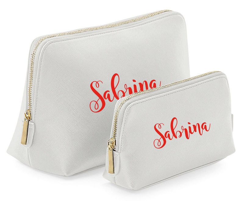 May include: Two white zippered cosmetic bags with gold zippers. The larger bag has the name "Sabrina" written in red cursive. The smaller bag has the same name written in red cursive.