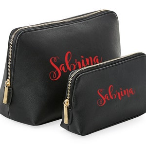 May include: Two black zippered cosmetic bags with gold zippers. The larger bag has the name "Sabrina" in red cursive lettering. The smaller bag has the same name in red cursive lettering.