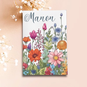 Può includere: Una stampa su tela rettangolare con il nome "Manon" in corsivo sopra una vivace illustrazione floreale. L'opera presenta una varietà di fiori colorati, tra cui papaveri arancioni, dalie rosa e fiordalisi blu, su uno sfondo bianco.