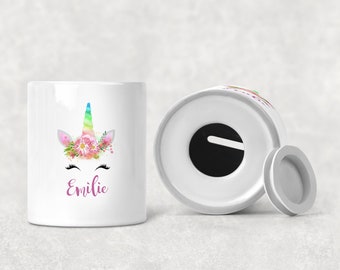 Può includere: Salvadanaio in ceramica bianca a forma di unicorno con un corno arcobaleno e fiori rosa. Il salvadanaio ha una fessura nella parte superiore e un coperchio rimovibile. Il nome "Emilie" è scritto in rosa sul davanti.