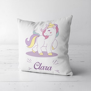 Peut inclure: Un oreiller blanc avec une illustration de licorne colorée et le nom "Clara" imprimé en violet.