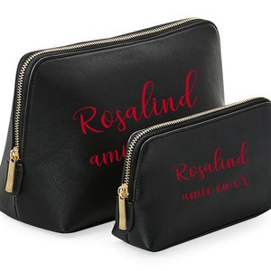 May include: Two black zippered makeup bags with gold zippers. The larger bag has the name "Rosalind" written in red cursive lettering. The smaller bag has the name "Rosalind" written in red cursive lettering and the words "amie coeur" written in red cursive lettering below.