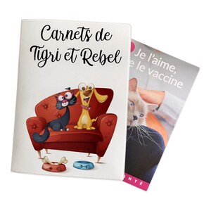 May include: A white booklet with the text "Carnets de Tigri et Rebel" and an illustration of a black cat and a yellow dog sitting on a red couch. The booklet is partially covered by a white sheet of paper with the text "Je l'aime, le vaccine" in black.