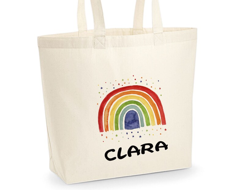 May include: A white canvas tote bag with a rainbow and stars design. The name "CLARA" is printed in black below the rainbow.