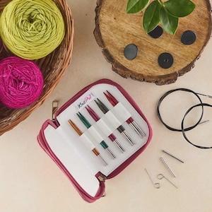 Könnte beinhalten: Ein weinrotes Etui mit Reißverschluss und KnitPro-Branding, geöffnet, um ein Set bunter Stricknadeln zu zeigen. Zwei Knäuel Garn, eines grün und eines rosa, liegen in einem Weidenkorb. Zusätzliche Strickwerkzeuge und eine Holzscheibe sind ebenfalls sichtbar.