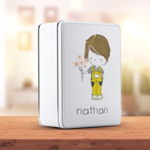 Op de afbeelding: Witte metalen doos met een cartoonillustratie van een jongen die bloemen vasthoudt. De jongen draagt een gele overall en de doos heeft de naam "nathan" onderaan gedrukt.