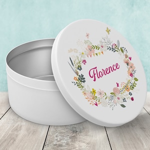 Peut inclure: Une boîte ronde blanche avec un couvercle orné d'une couronne florale. Le mot "Florence" est écrit en cursive à l'intérieur de la couronne. La boîte est ouverte, révélant un intérieur argenté, et posée sur une surface en bois.