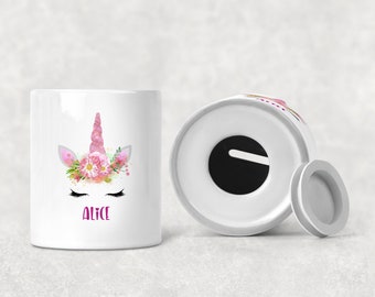 Può includere: Salvadanaio in ceramica bianca con un design a forma di viso di unicorno. L'unicorno ha fiori rosa e un corno rosa. Il salvadanaio ha una fessura nella parte superiore e un coperchio rimovibile. Il nome "Alice" è scritto in lettere rosa sul davanti.