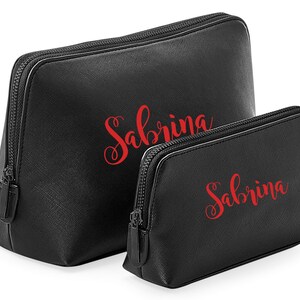May include: Two black cosmetic bags, each with the name "Sabrina" in red script. The bags have a textured finish and black zippers. The larger bag is taller than the smaller one. Designed for makeup and beauty products.