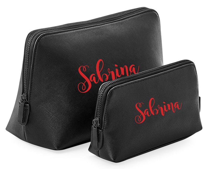 May include: Two black cosmetic bags, each with the name "Sabrina" in red script. The bags have a textured finish and black zippers. The larger bag is taller than the smaller one. Designed for makeup and beauty products.