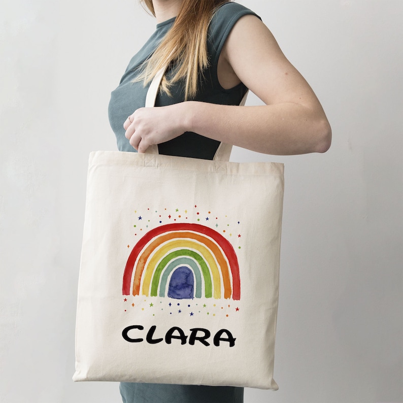 May include: A white canvas tote bag with a rainbow design and the name "CLARA" printed in black. The rainbow is made of watercolor-style stripes in red, orange, yellow, green, blue, and purple. There are small white stars scattered around the rainbow.