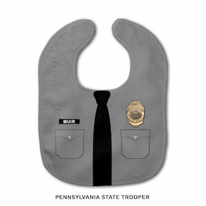 Könnte beinhalten: Ein graues Baby-Lätzchen im Design einer Uniform der Pennsylvania State Police. Es zeigt eine schwarze Krawatte, zwei Brusttaschen, ein goldfarbenes Abzeichen und ein schwarzes Namensschild mit dem weißen Text MUIR. Darunter steht PENNSYLVANIA STATE TROOPER.