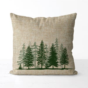 May include: A square decorative pillow with a natural linen-textured cover. The pillow features a forest scene with dark green pine trees silhouetted against a lighter green background. The trees are arranged in a row.