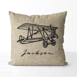 May include: A square pillow with a linen texture features a black line drawing of a vintage biplane. The name "Jackson" is written in cursive script below the plane. The pillow is a neutral beige color.