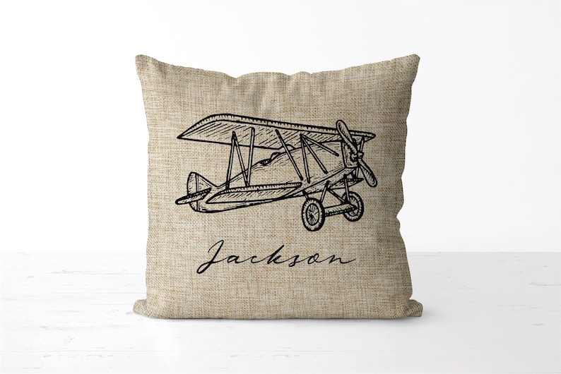May include: A square pillow with a linen texture features a black line drawing of a vintage biplane. The name "Jackson" is written in cursive script below the plane. The pillow is a neutral beige color.