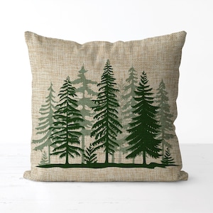 May include: A decorative pillow with a natural linen-textured cover. The pillow features a forest scene with several evergreen trees in varying shades of green. The trees are silhouetted against the light background, creating a nature-inspired design.