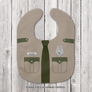 Può includere: Una bavaglina color marrone chiaro disegnata per sembrare un'uniforme della Tennessee Highway Patrol. Presenta una cravatta verde scuro, due tasche sul petto, un distintivo argentato e una targhetta con il nome RAINS. Il testo TENNESSEE HIGHWAY PATROL è stampato in basso.
