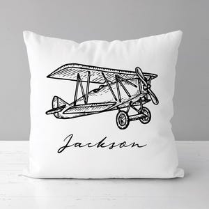 May include: White square pillow featuring a black and white line drawing of a biplane. The biplane has a propeller, wheels, and a detailed wing design. The name "Jackson" is written in cursive below the plane.