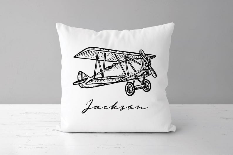 May include: White square pillow featuring a black and white line drawing of a biplane. The biplane has a propeller, wheels, and a detailed wing design. The name "Jackson" is written in cursive below the plane.