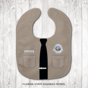 May include: A tan baby bib designed to resemble a Florida State Highway Patrol uniform. It features a black necktie, two chest pockets, a name tag reading KASHER, and a circular badge with the text HIGHWAY PATROL TROOPER and FLORIDA STATE HIGHWAY PATROL.