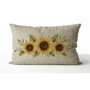 May include: Rectangular beige pillow with a watercolor design of three sunflowers and green leaves. The sunflowers have yellow petals and brown centers. The pillow has a textured, burlap-like appearance.