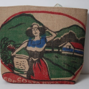 May include: A brown burlap tote bag with a colorful illustration of a woman in a red dress and a blue top, holding a basket. The illustration is set against a green landscape with a small house in the background. The text "COSTA RICA" is printed on the bottom of the bag.