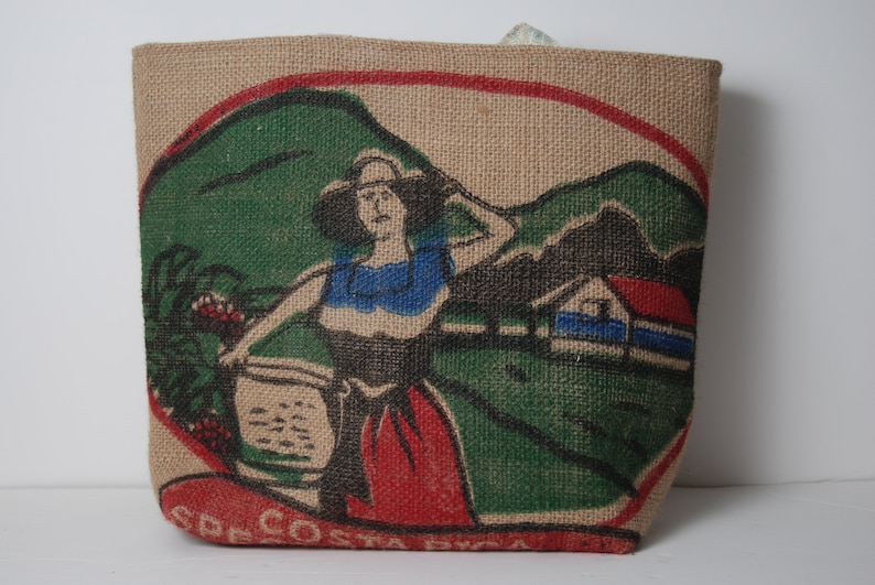 May include: A brown burlap tote bag with a colorful illustration of a woman in a red dress and a blue top, holding a basket. The illustration is set against a green landscape with a small house in the background. The text "COSTA RICA" is printed on the bottom of the bag.