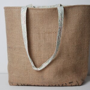 May include: A large brown burlap tote bag with fabric handles featuring a floral pattern. The bag is made of natural fibers and has a rustic look.