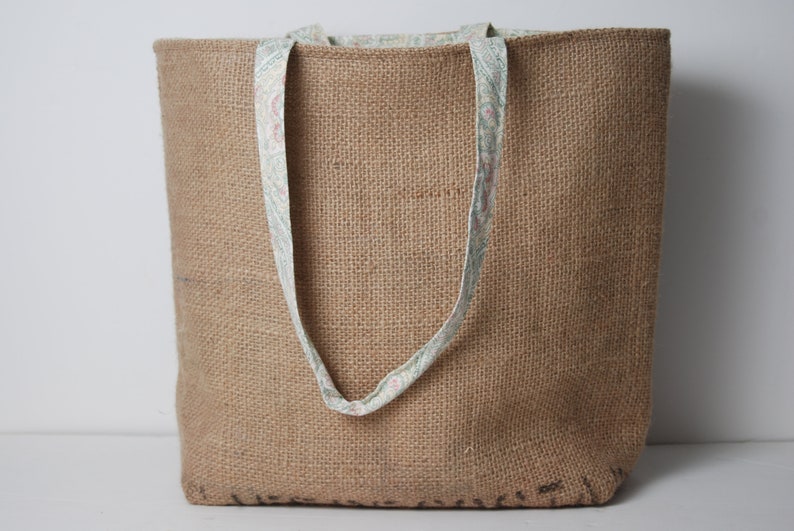 May include: A large brown burlap tote bag with fabric handles featuring a floral pattern. The bag is made of natural fibers and has a rustic look.