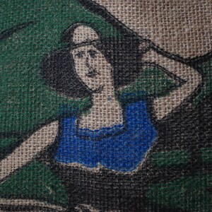 May include: A burlap fabric with a painted image of a woman wearing a blue top. The woman has dark hair and is looking to the right. The background is green and brown.