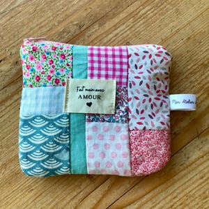 Peut inclure: Une petite pochette patchwork avec une fermeture éclair rose. Elle est composée de différents motifs de tissus, dont des motifs floraux, vichy et à pois. Une étiquette indique "Fait main avec AMOUR" avec un cœur.