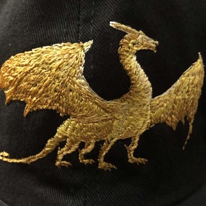 Puede incluir: Una gorra de béisbol negra con un diseño de dragón bordado en oro.