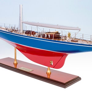 Può includere: Un modello dettagliato di una barca a vela vintage con scafo blu e rosso, montato su una base in legno. Il modello presenta dettagli intricati, tra cui un albero, sartiame e una piccola vela.