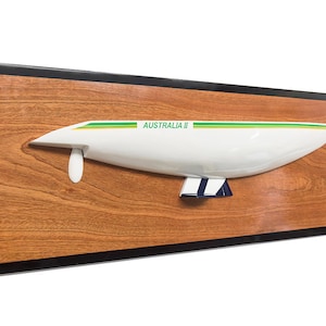 Könnte beinhalten: Ein weißes Modell der Yacht Australia II mit grünen und gelben Streifen, montiert auf einer Holzplatte mit schwarzem Rahmen. Die Platte hat eine kleine goldene Plakette mit dem Text "AUSTRALIA II" darauf.