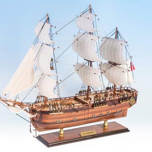 May include: A detailed wooden model of a tall ship with white sails and rigging. The ship has a brown hull and deck, with cannons visible along the sides. It is mounted on a wooden base with a nameplate.