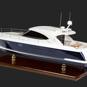 May include: A white and blue yacht model with a wooden base. The yacht has a white cabin with a dark blue hull and a white stripe along the bottom. The yacht is on a wooden base with gold-coloured supports.