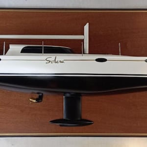 Könnte beinhalten: Ein weißes und schwarzes Modell eines Segelboots mit dem Namen "Solera" an der Seite, das in einem Holzrahmen ausgestellt ist.