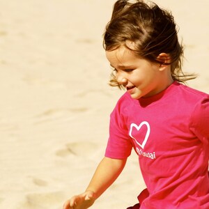May include: A young girl with brown hair is wearing a pink t-shirt with a white heart and the word "Maui" on it. She is smiling and looking down.