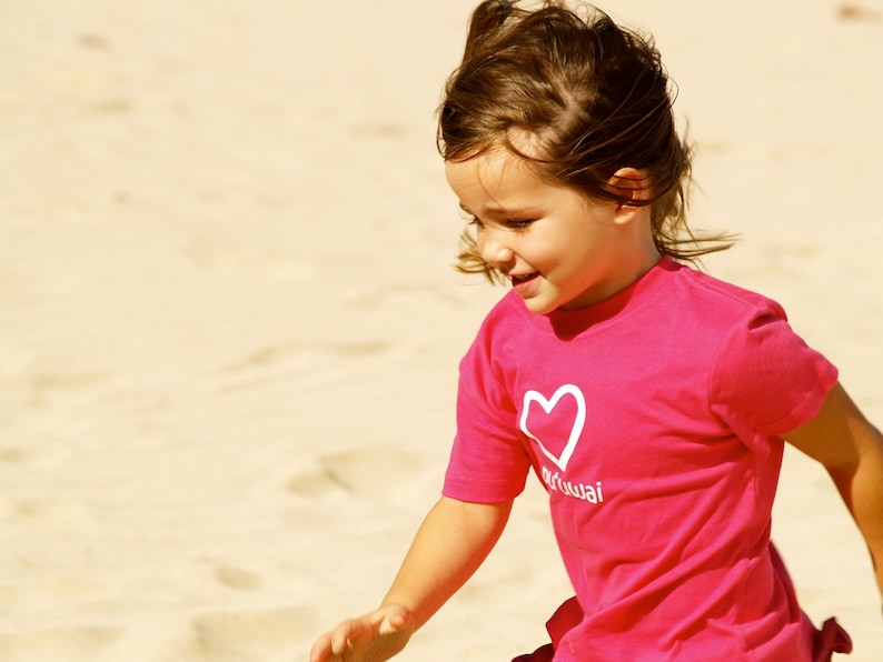 May include: A young girl with brown hair is wearing a pink t-shirt with a white heart and the word "Maui" on it. She is smiling and looking down.
