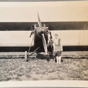 May include: Black and white photograph of a biplane with a person standing in front of it. The biplane has two wings, a propeller, and a person standing in front of it. The person is wearing a vest and shorts. The photograph is taken outdoors.